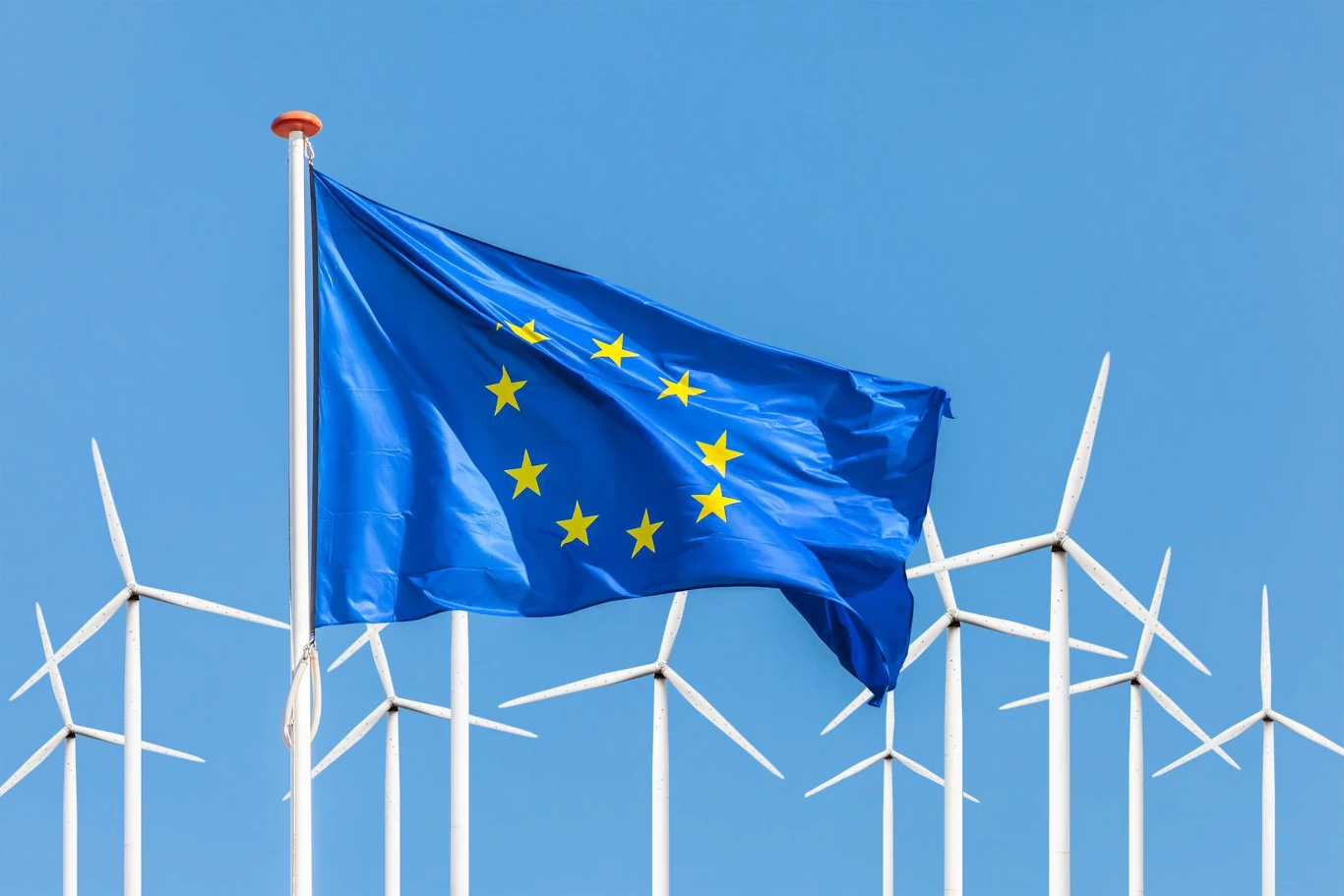 Europa na drodze ku energetycznej niezależności Flaga Unii Europejskiej powiewająca na tle licznych białych turbin wiatrowych na niebieskim niebie.