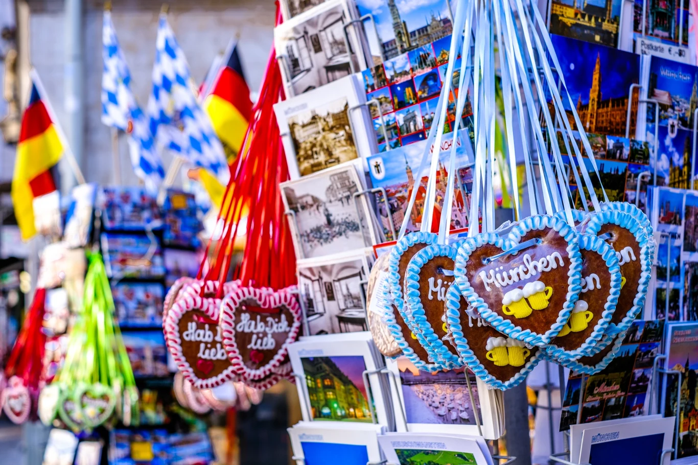 Kolorowe pierniki w kształcie serca z napisem oraz liczne widokówki z wizerunkami miast, na tle flag bawarskiej i niemieckiej, które sugerują stoisko pamiątkowe w miejscu turystycznym.