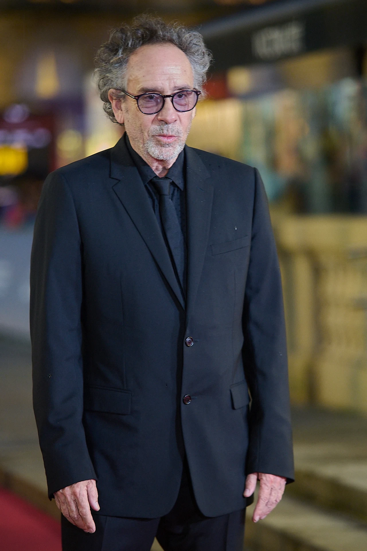 Tim Burton Mężczyzna w średnim wieku ubrany w elegancki czarny garnitur i okulary stoi na tle rozmytego, miejskiego pejzażu z elementem czerwonego dywanu.