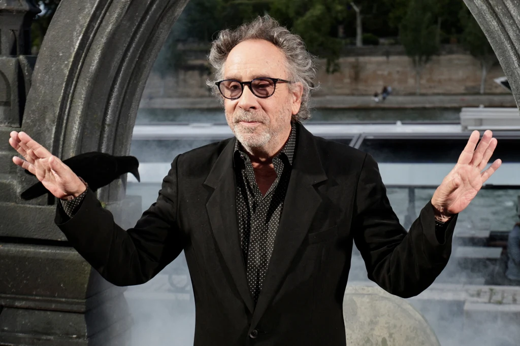 Tim Burton Mężczyzna w średnim wieku w okularach i ciemnym garniturze stoi z uniesionymi rękami, w tle kamienna architektura, elementy dymu i kruk na łuku po lewej stronie.