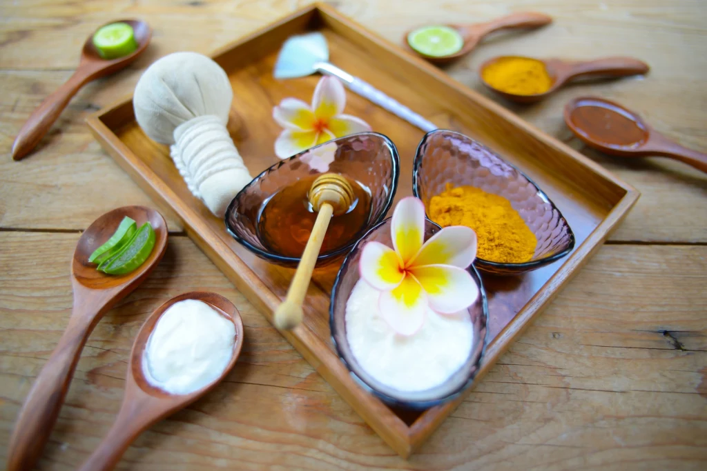 Naturalne składniki w tajskiej pielęgnacji Drewniana taca z trzema szklanymi miseczkami wypełnionymi miodem, białym kremem i sproszkowaną kurkumą, ozdobiona kwiatami plumerii, otoczona drewnianymi łyżkami z naturalnymi składnikami i przyborami kosmetycznymi.