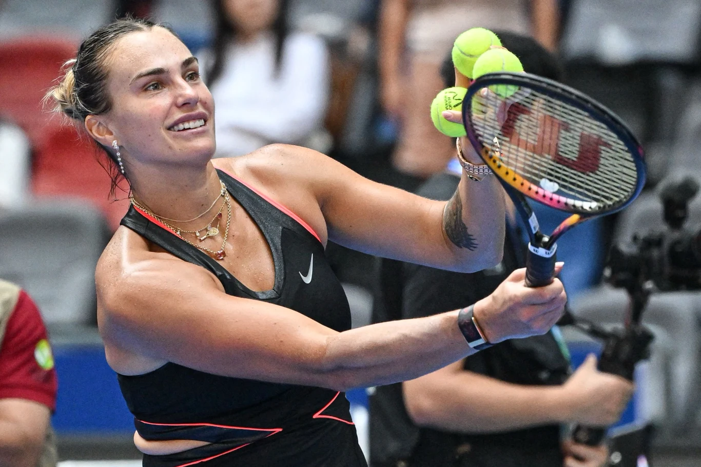 Zadowolona tenisistka w sportowym stroju odbija piłki tenisowe rakietą na hali sportowej podczas wydarzenia z udziałem publiczności.