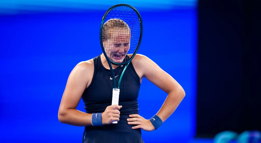 Mirra Andriejewa Zawodniczka tenisowa w ciemnoniebieskim stroju sportowym trzymająca rakietę przed twarzą, wyrażająca silne emocje w trakcie meczu na korcie o intensywnie niebieskim tle.