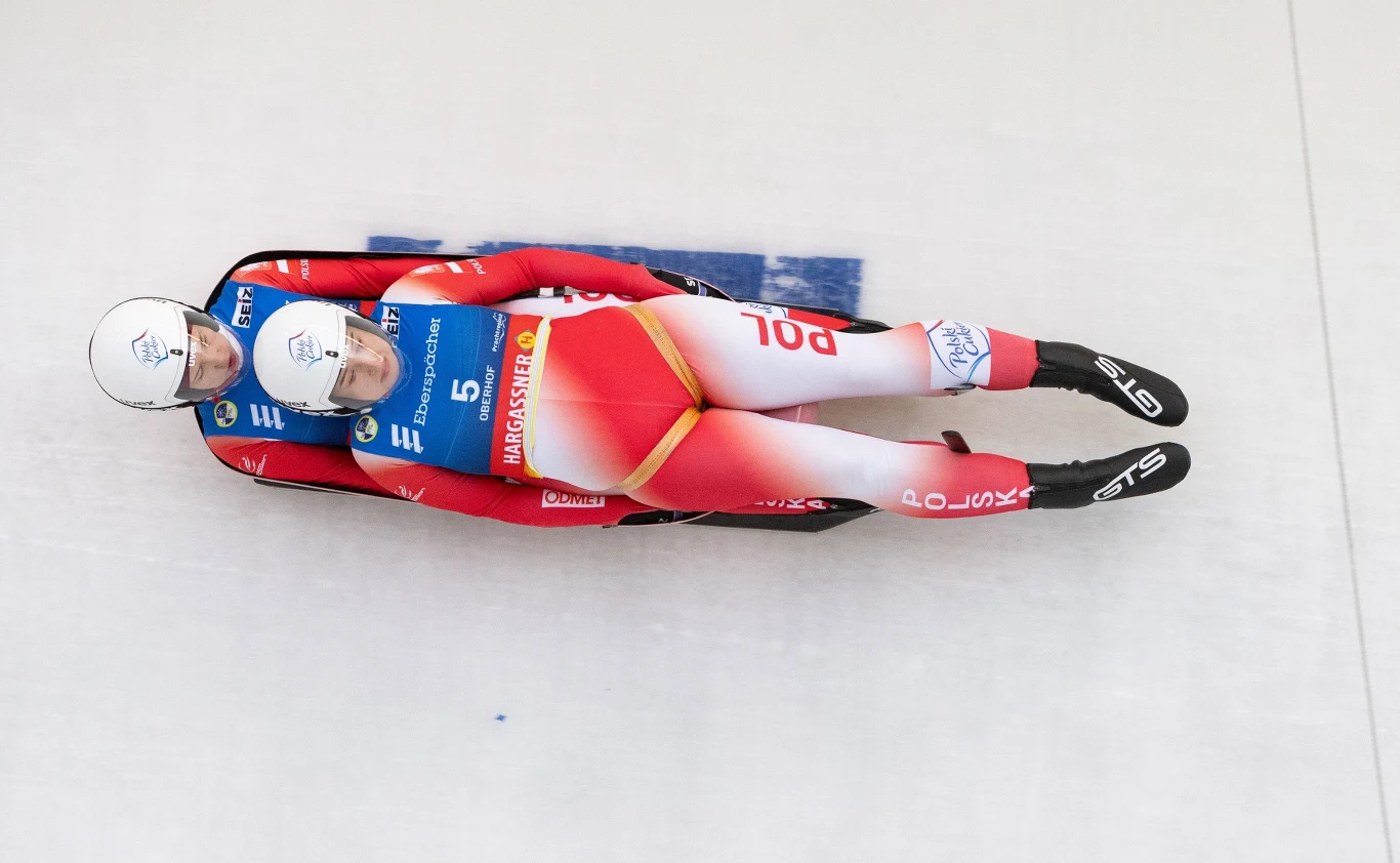 Nasze saneczkarki powalczą o czołowe lokaty na zimowych igrzyskach olimpijskich 2026 Dwóch sportowców w kaskach i kombinezonach startowych leży na saneczkach w pozycji aerodynamicznej podczas konkurencji saneczkarskiej na lodowym torze