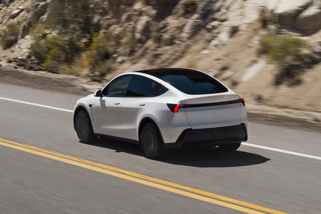 Tesla Model Y Standard Biały samochód elektryczny typu SUV jedzie po asfaltowej drodze wśród skalistego krajobrazu, auto widoczne z tyłu, dynamiczny ruch podkreślony rozmytym tłem.