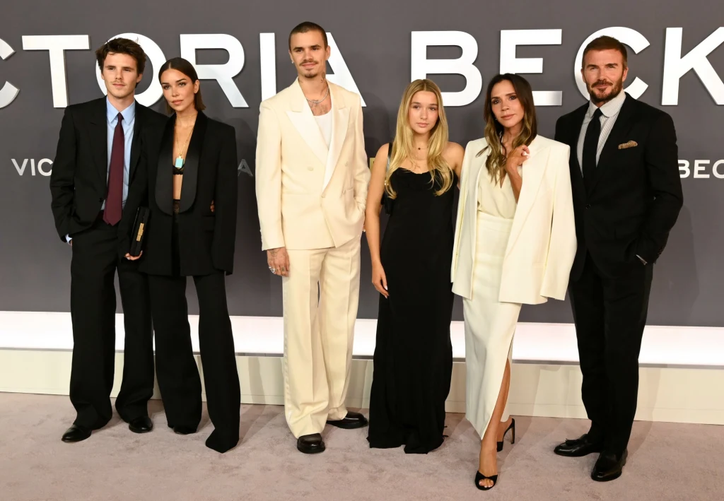 Premiera serialu Victoria Beckham Sześć elegancko ubranych osób stoi razem na tle ściany z dużym napisem, część jest w jasnych garniturach i sukniach, uśmiechają się i pozują do zdjęcia na oficjalnym wydarzeniu.