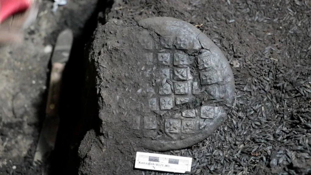 Spalony, okrągły bochenek chleba z widocznymi odciśniętymi kwadratami na powierzchni, leżący w ciemnej ziemi obok miarki archeologicznej