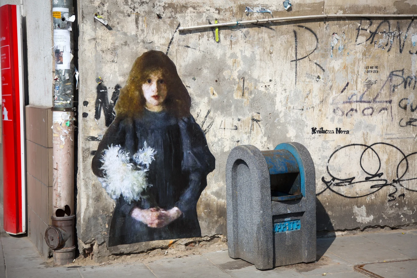 Portret młodej kobiety w ciemnej sukni namalowany na zniszczonym murze, obok którego stoi szara uliczna kosz na śmieci, ściana jest pokryta graffiti i śladami wandalizmu.