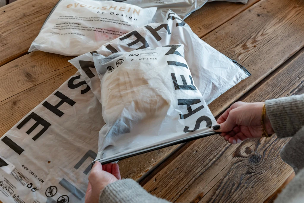 Firma Shein planuje również otwarcie sklepów w popularnych domach towarowych Galeries Lafayette Ręce rozpakowujące przesyłkę z ubraniami w plastikowym opakowaniu z logo SHEIN położoną na drewnianym stole, w tle widoczne inne opakowania z tej samej marki.