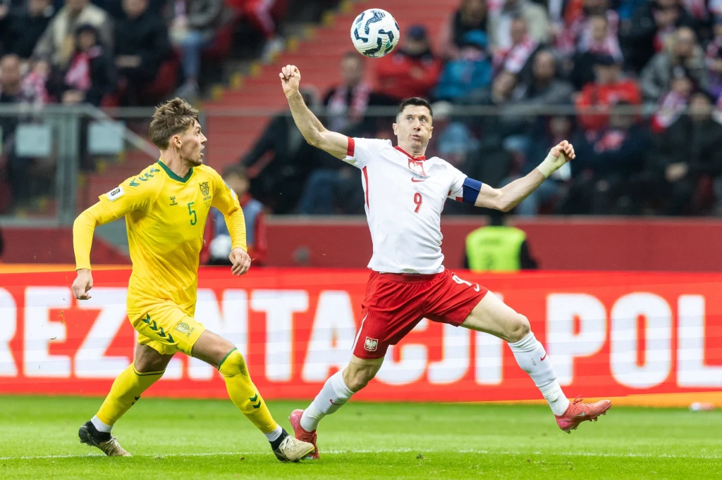 Dwóch piłkarzy podczas dynamicznej akcji na boisku, zawodnik w żółtym stroju biegnie tuż obok piłkarza w białej koszulce z numerem 9, który odbiera piłkę w powietrzu; w tle widoczna publiczność stadionowa.