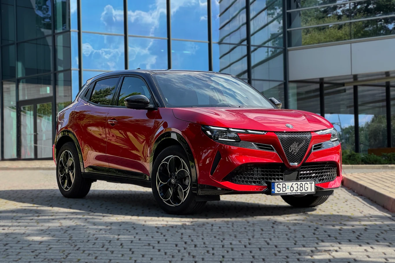 Alfa Romeo Junior, niezależnie od wersji, produkowana jest w Tychach Czerwony samochód Alfa Romeo Tonale zaparkowany na brukowanym placu przed nowoczesnym szklanym budynkiem, w tle odbijające się w szybach drzewa i niebo.