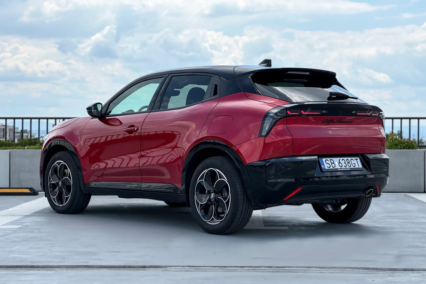 Bliźniacze modele Alfy Junior to Fiat 600 i Jeep Avenger Czerwony crossover Alfa Romeo Tonale zaparkowany na otwartym parkingu, widok od tyłu, charakterystyczne tylne światła LED, czarne wykończenia dachu, sportowe felgi, widoczna tablica rejestracyjna.