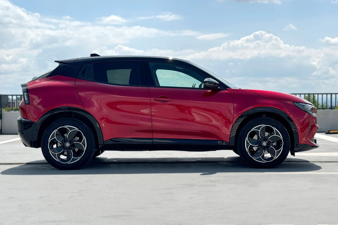 Chociaż technicznie Fiat, Alfa i Jeep to te same modele, stylizacyjnie są zupełnie inne Czerwony, nowoczesny samochód typu crossover zaparkowany na otwartym parkingu z widokiem na niebo i chmury w tle.