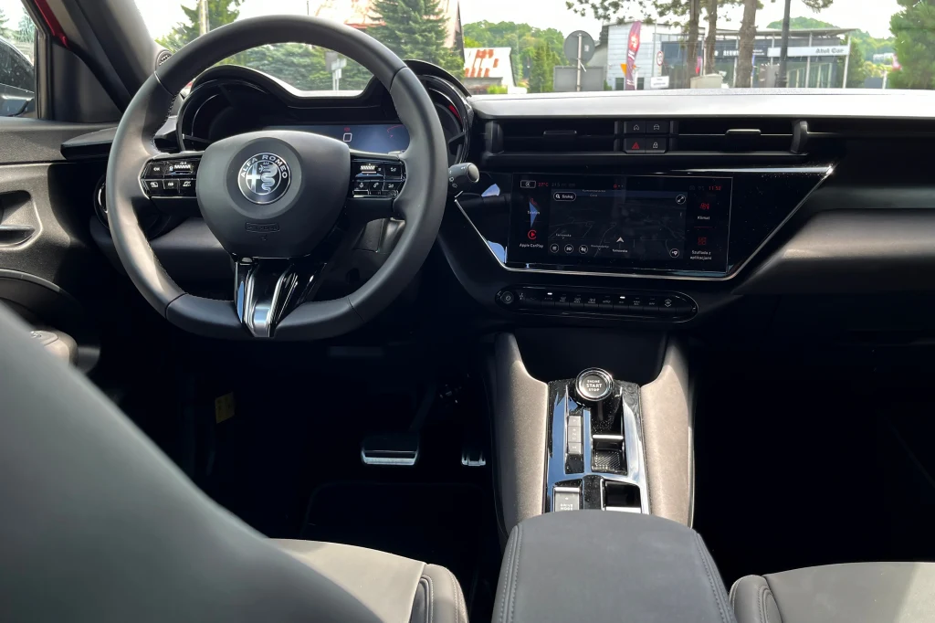 Kokpit nowoczesnego samochodu marki Alfa Romeo z dużym, centralnym ekranem dotykowym, sportową kierownicą oraz elegancką konsolą środkową, wykończoną na wysoki połysk.