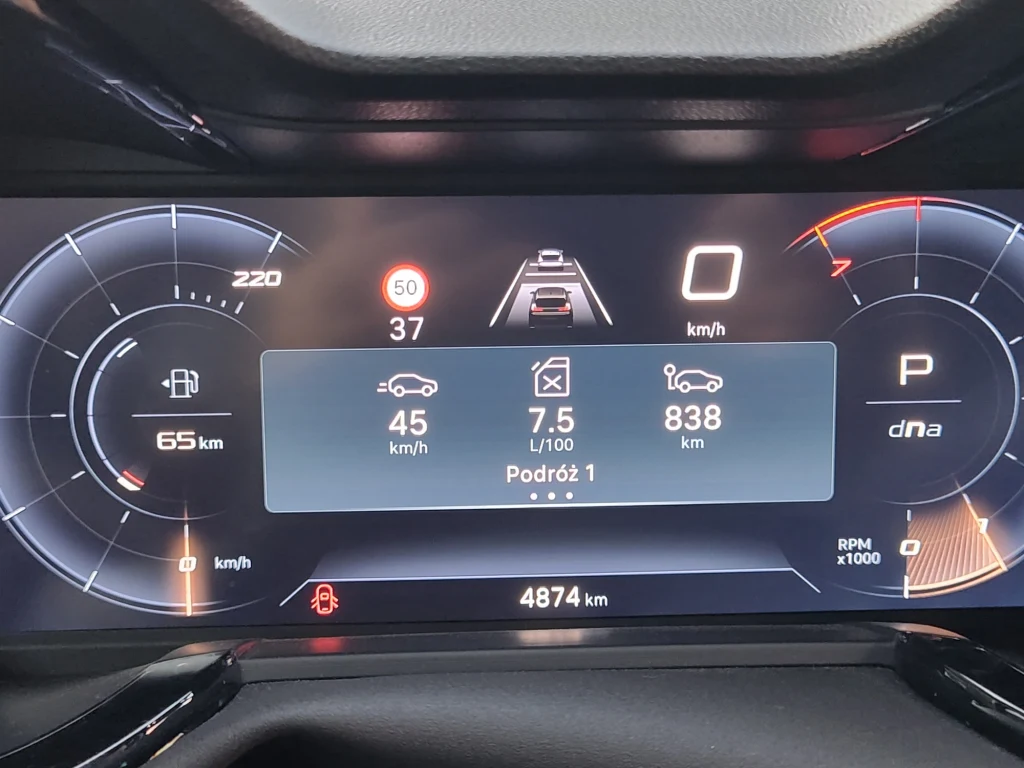 Cyfrowy wyświetlacz deski rozdzielczej samochodu z czytelnymi wskaźnikami prędkości, poziomu paliwa, liczby przejechanych kilometrów oraz informacjami dotyczącymi zużycia paliwa i zasięgu. Centralnie znajdują się dane o średniej prędkości, spalaniu i m...