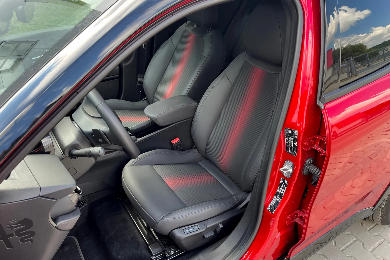 Fotele Alfy Junior są wygodne Wnętrze nowoczesnego samochodu z przodu, uchylone czerwone drzwi, czarne fotele z czerwonym akcentem i widoczna deska rozdzielcza.