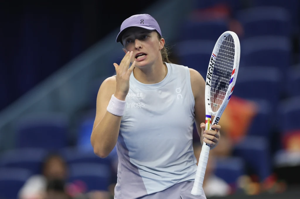 Tenisistka ubrana w sportowy strój, trzymająca rakietę tenisową, wykonuje gest ręką przy ustach, będąc na korcie podczas rozgrywki.