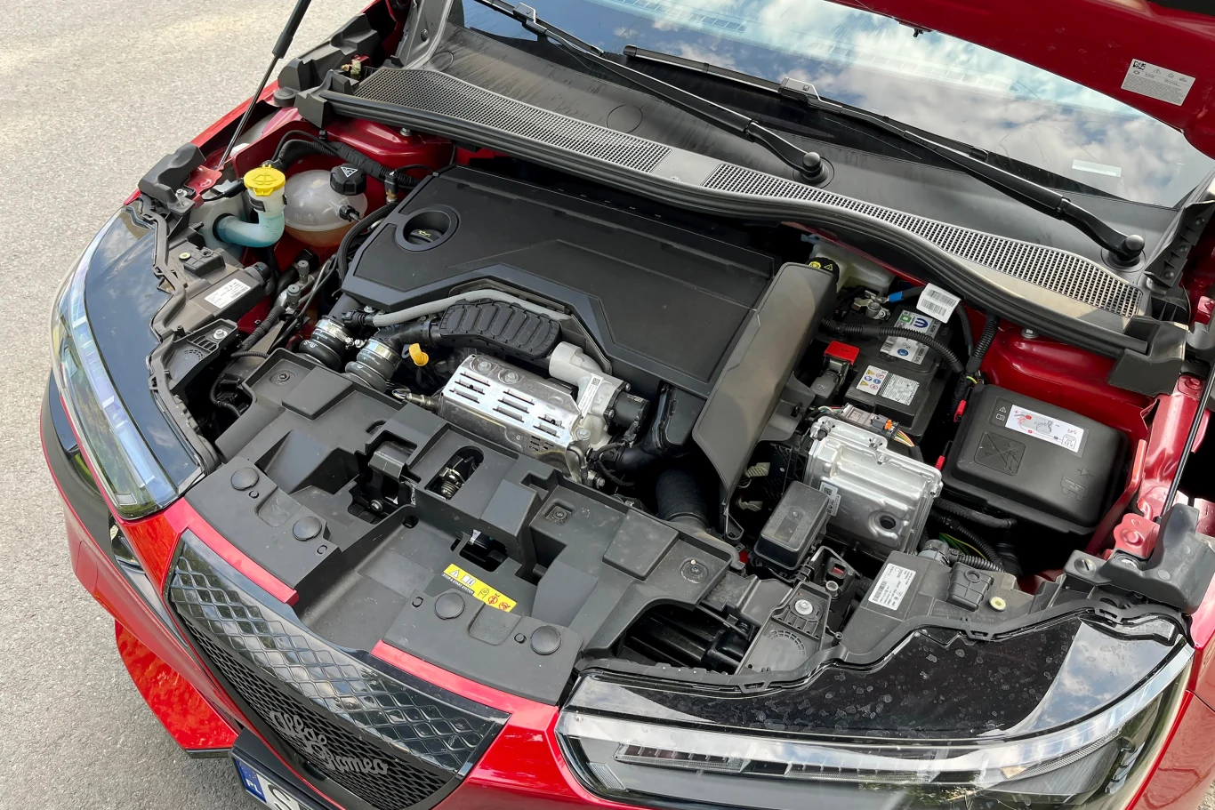 Napęd Juniora Q4 składa się z silnika elektrycznego zintegrowanego z przekładnią dwusprzęgłową, silnika spalinowego 1.2 i silnika elektrycznego przy tylnej osi Otwarta maska czerwonego samochodu, pod którą widoczny jest silnik i inne elementy mechaniczne, w tym akumulator, zbiornik płynów i przewody instalacji elektrycznej.