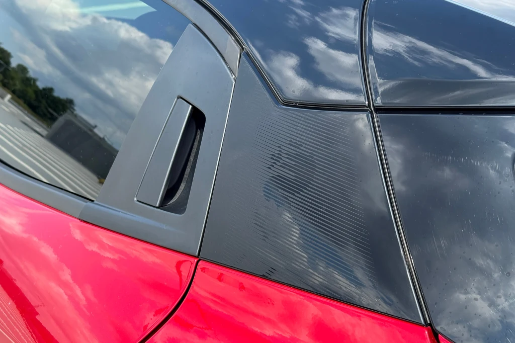 Fragment nadwozia samochodu w kolorze czerwonym z czarnym dachem, na szybie odbijają się chmury, widoczne są detale karoserii i linie podziału lakieru.