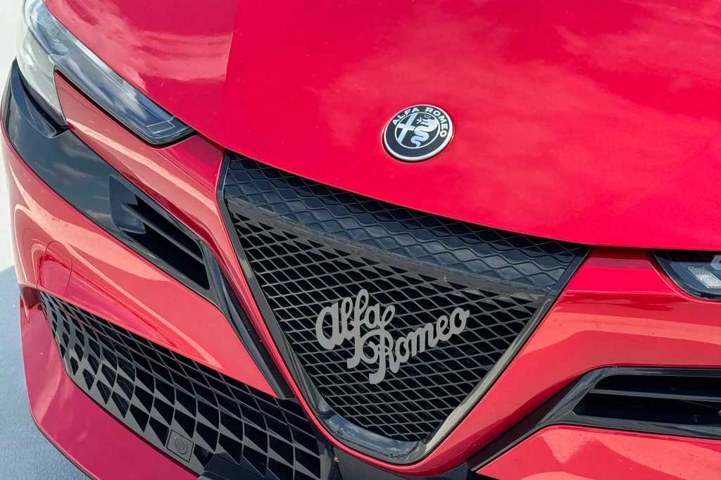 Czerwony przód samochodu Alfa Romeo z charakterystycznym logo marki oraz wyrazistym grillem o geometrycznym wzorze.