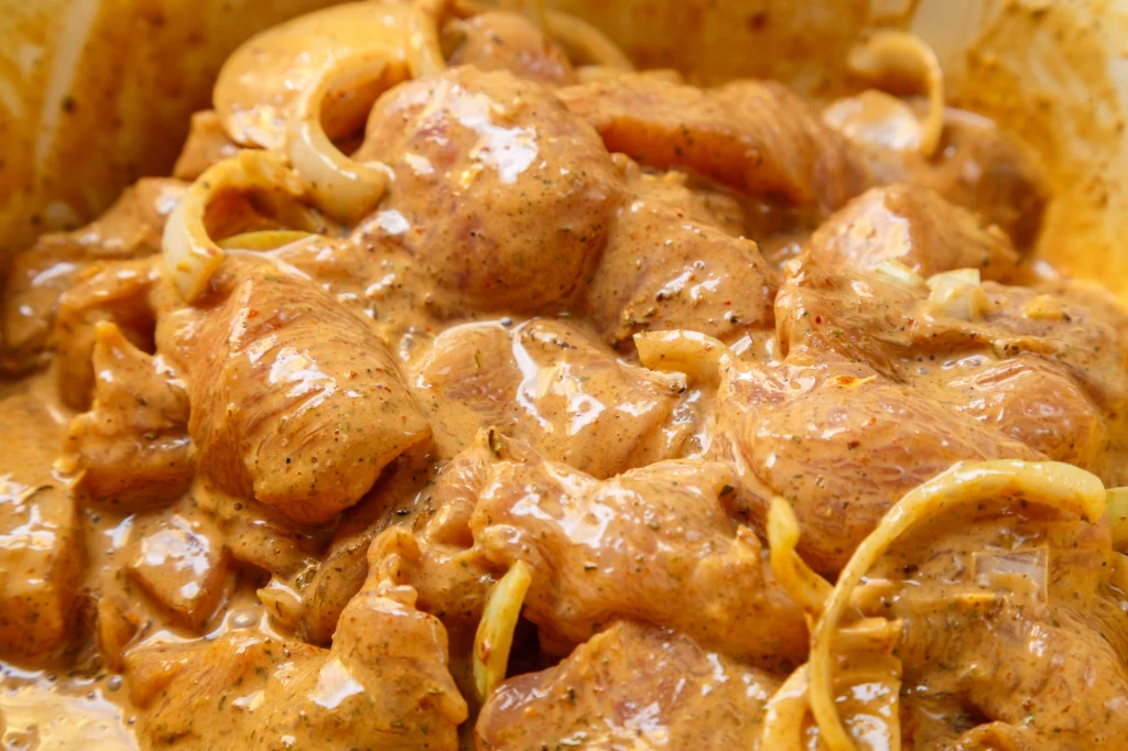 Mięso na gulasz możesz zamarynować nawet dzień wcześniej i zostawić w lodówce na całą noc Kawałki surowego mięsa zatopione w marynacie z przyprawami i plasterkami cebuli, całość pokryta kremowym sosem o intensywnym, żółto-brązowym kolorze.