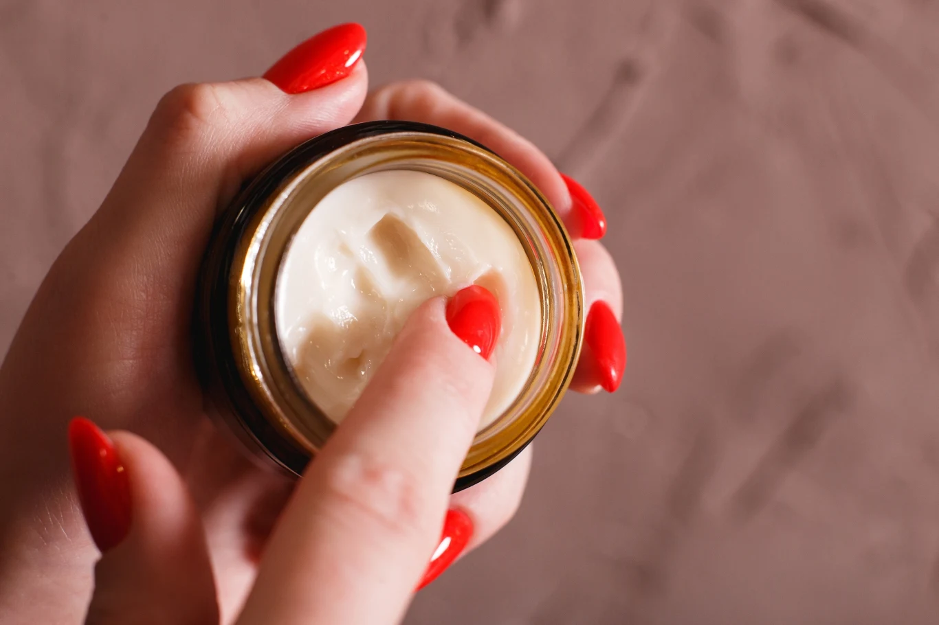 Ręka z czerwonymi paznokciami nabiera krem z brązowego słoiczka na tle jasnobrązowej powierzchni.
