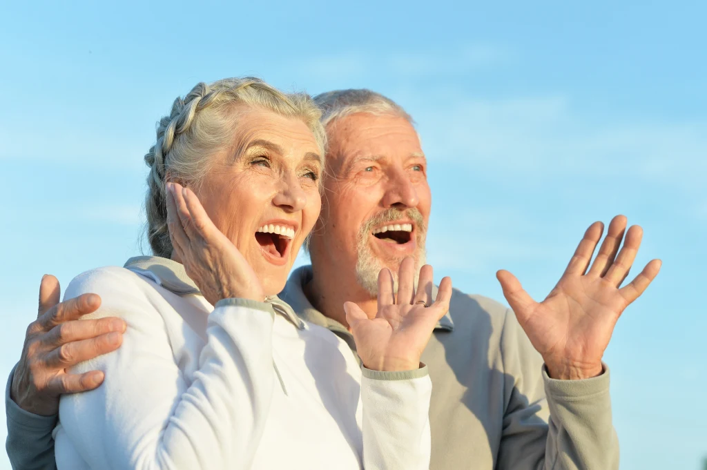 Pogoda ducha i otwartość to jeden z przepisów na długie życie w zdrowiu Starsza kobieta i mężczyzna z szerokimi uśmiechami, stoją objęci i zaskoczeni, gestykulując dłoniami na tle jasnego nieba.