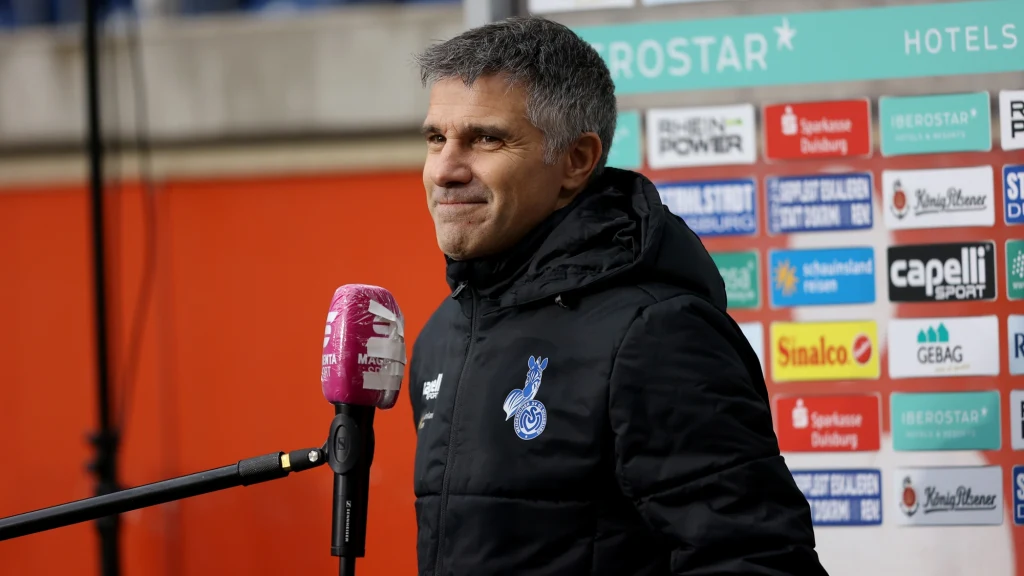 Gino Lettieri Mężczyzna w czarnej kurtce sportowej udziela wywiadu, stojąc przy mikrofonie, w tle widoczne tło z logotypami sponsorów sportowych.