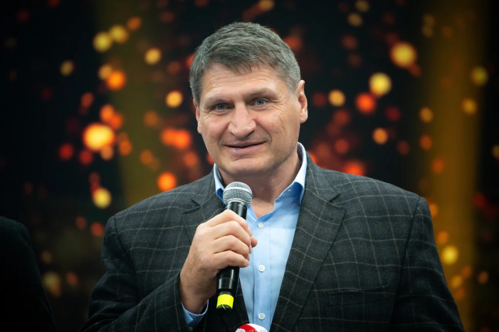 Mężczyzna w średnim wieku w marynarce w kratę i niebieskiej koszuli trzyma mikrofon, stoi na scenie z rozmytym tłem pełnym świetlistych punktów.