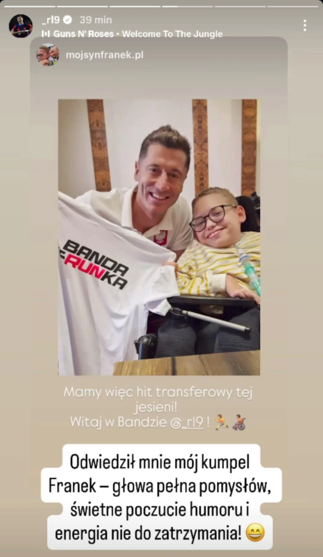 Robert Lewandowski wraz z "kumplem" Uśmiechnięty mężczyzna trzyma koszulkę z napisem 'Banda Franka' obok siedzącego na wózku chłopca w okularach, oboje wyglądają na zadowolonych i pełnych energii, tło stanowi wnętrze mieszkania.