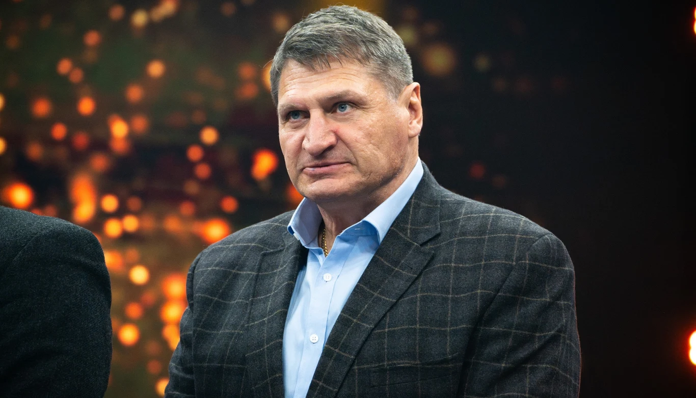 Mężczyzna w średnim wieku w eleganckiej marynarce w kratę oraz jasnoniebieskiej koszuli stoi na tle ciemnego tła z żółtopomarańczowymi światłami bokeh.