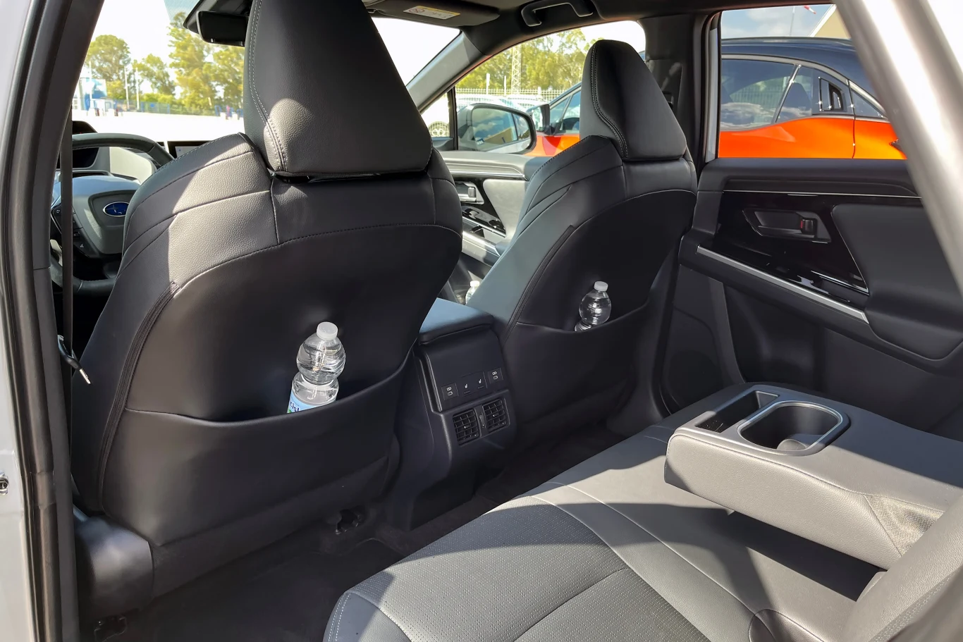 Wnętrze samochodu osobowego z widocznymi tylnymi siedzeniami, butelkami wody w kieszeniach siedzeń oraz środkowym podłokietnikiem. Na zewnątrz widoczne inne zaparkowane pojazdy.