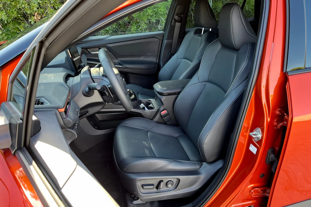 Przestronne wnętrze samochodu z widocznymi dwoma czarnymi, skórzanymi fotelami przednimi, kierownicą, deską rozdzielczą i konsolą środkową w nowoczesnym wykończeniu, drzwi od strony kierowcy otwarte, karoseria w kolorze pomarańczowym.