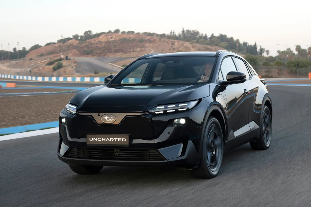 Czarny, nowoczesny samochód elektryczny jedzie po asfaltowym torze wyścigowym, otoczonym barierkami i wzgórzami w tle. Na masce i tablicy rejestracyjnej widoczne są charakterystyczne emblematy i nazwa modelu.