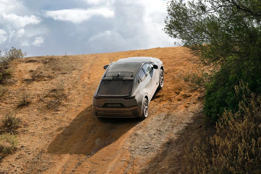 Samochód terenowy pokonuje strome, piaszczyste wzniesienie, otoczony przez niską roślinność, widoczny jest kurz unoszący się spod kół oraz niewielkie chmury na niebie.