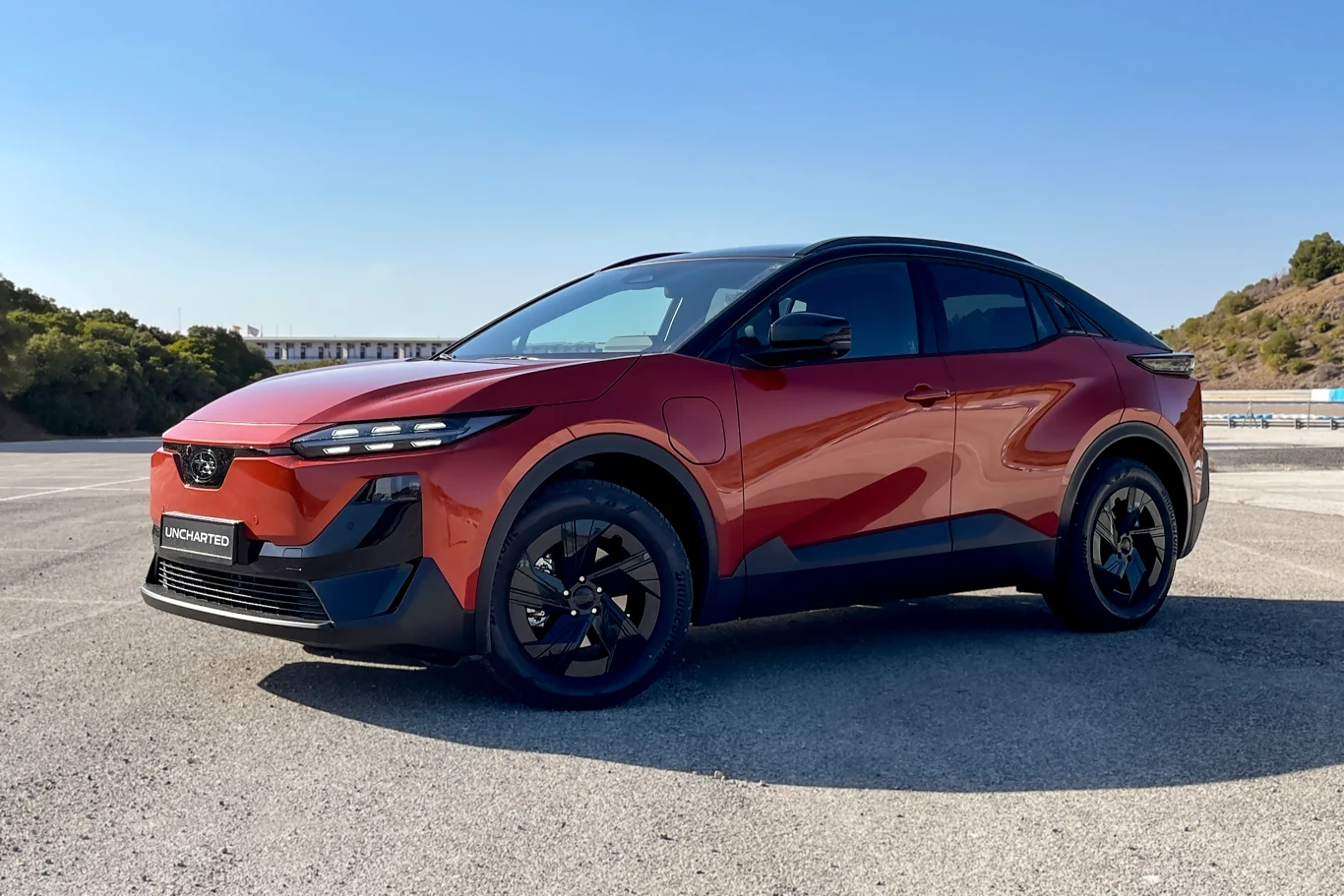 Czerwony samochód osobowy typu SUV z czarnymi elementami nadwozia stoi zaparkowany na otwartym placu asfaltowym, w tle widoczne są drzewa i nasłonecznione wzgórza.