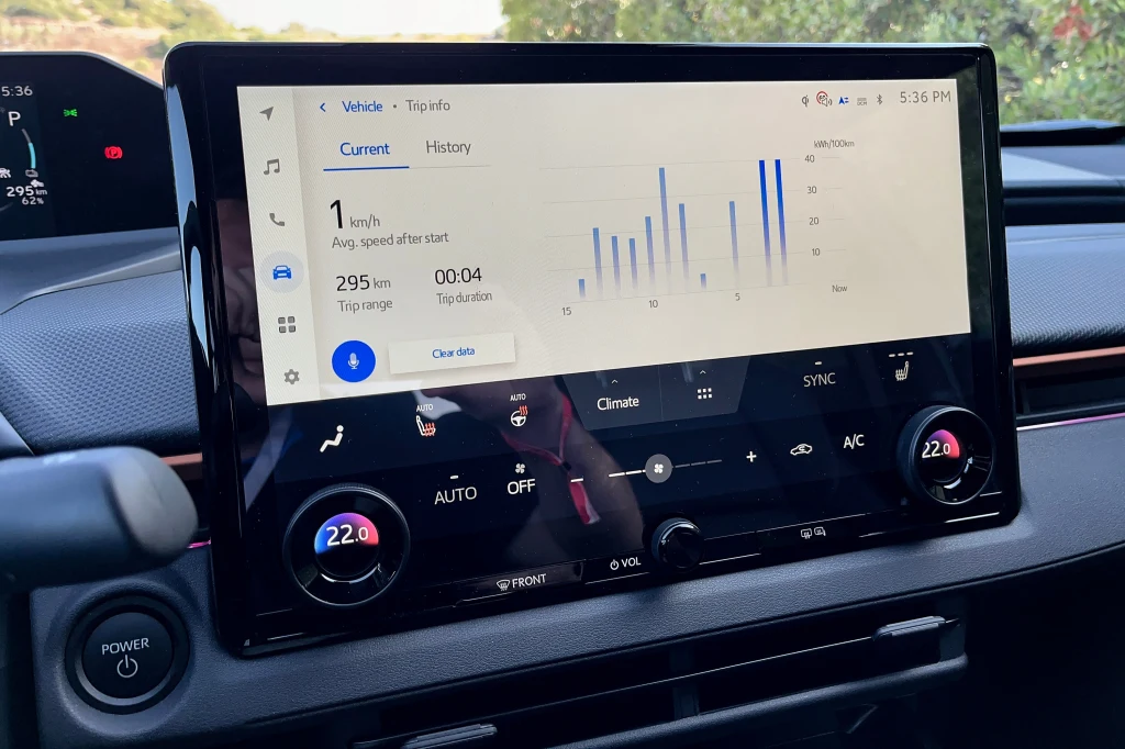 Centralny ekran systemu multimedialnego samochodu wyświetlający statystyki dotyczące podróży, z informacjami o średniej prędkości, dystansie oraz szczegółowym wykresem, poniżej znajdują się cyfrowe sterowanie klimatyzacją i panel przycisków.