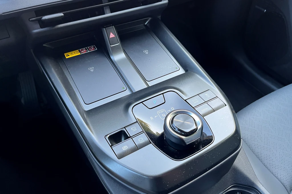 Konsola centralna samochodu z nowoczesnym, obrotowym panelem zmiany biegów, przyciskami funkcyjnymi oraz dwoma panelami dotykowymi umieszczonymi w pobliżu dźwigni.