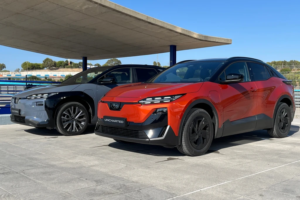 Dwa nowoczesne samochody elektryczne stojące obok siebie na otwartym placu, jeden w kolorze pomarańczowo-czarnym, drugi ciemnoszary, w tle nowoczesna betonowa konstrukcja dachu oraz fragment toru lub placu testowego.