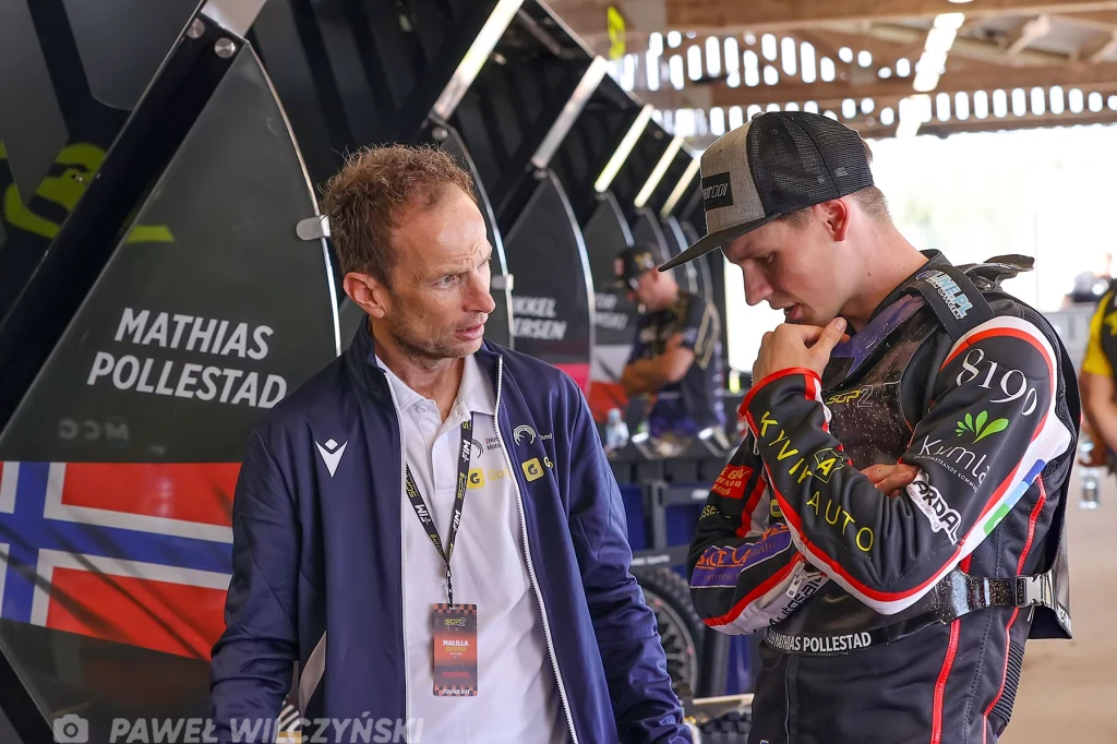 Dwóch mężczyzn rozmawia w warsztacie żużlowym, jeden ubrany w strój zawodnika z widocznymi logotypami sponsorów i flagą norweską w tle.
