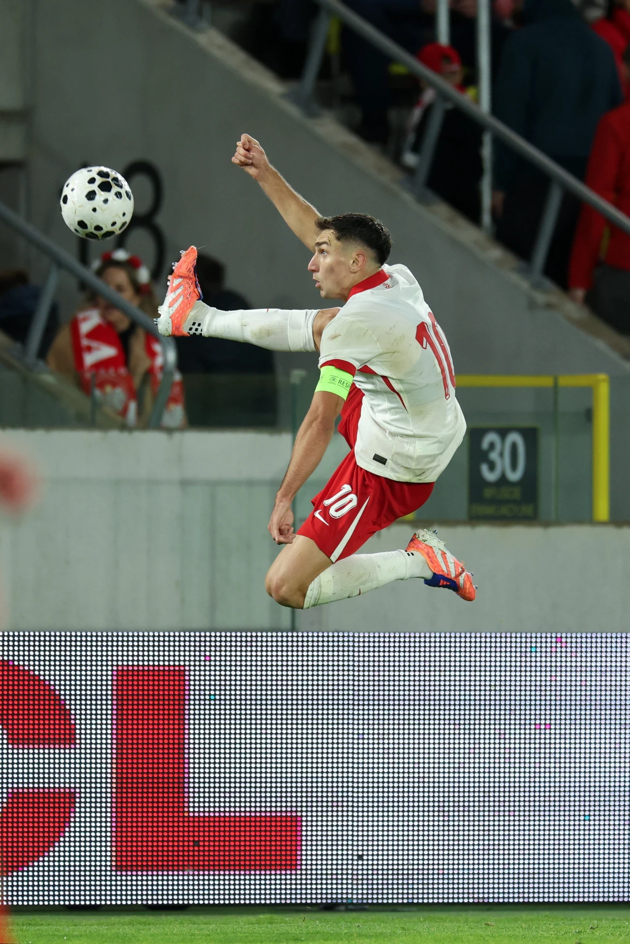 Piłkarz ubrany w biało-czerwony strój z numerem 10 i opaską kapitana wykonuje dynamiczny wyskok, próbując kopnąć piłkę w powietrzu podczas meczu, w tle trybuny z kibicami.