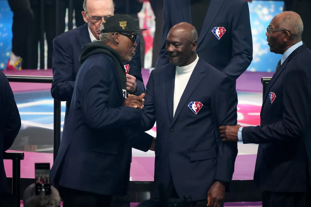 Dennis Rodman i Michael Jordan na gali z okazji 75-lecia istnienia NBA Grupa elegancko ubranych mężczyzn w marynarkach z naszywką logotypu NBA rozmawia ze sobą oraz uśmiecha się, jeden z nich wykonuje gest powitania dłonią.