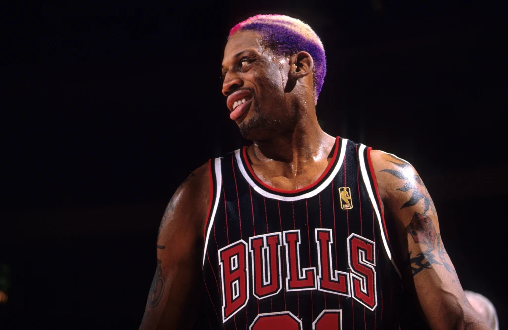 Dennis Rodman za czasów gry w Chicago Bulls Koszykarz drużyny Chicago Bulls w charakterystycznym stroju sportowym, z kolorowymi włosami i tatuażami na ramieniu, w trakcie meczu na ciemnym tle hali.