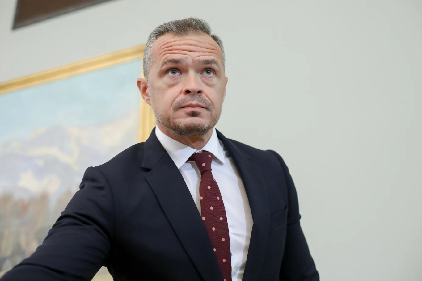 Sławomir Nowak Mężczyzna w eleganckim garniturze z czerwonym krawatem w białe kropki stoi na tle jasnego obrazu ściennego, z poważnym wyrazem twarzy patrzy lekko w górę.