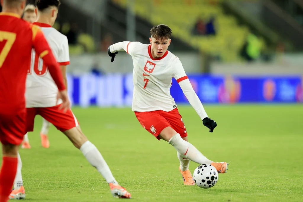 Oskar Pietuszewski zrobił kolejny krok w kierunku kadry A Piłkarz w białej koszulce z numerem 7 i godłem Polski kopie piłkę, otoczony przez zawodników przeciwnej drużyny w czerwonych strojach na stadionie podczas meczu piłki nożnej.