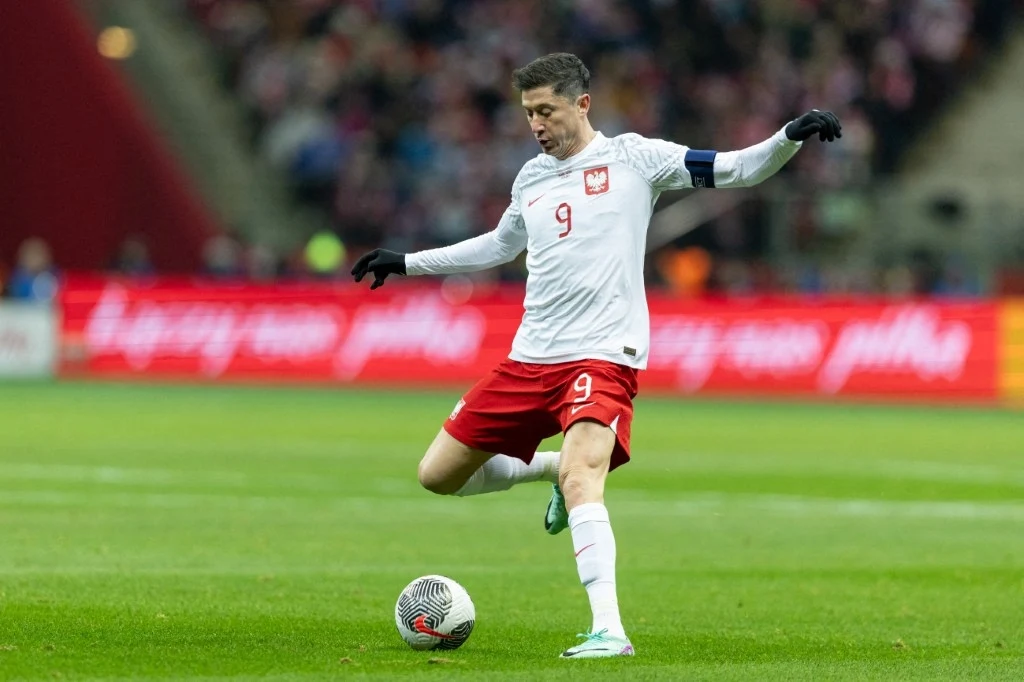 UEFA podjęła ważną decyzję przed meczem Litwa - Polska Piłkarz w białej koszulce z numerem 9 i czerwoną opaską kapitana wykonuje strzał podczas meczu na stadionie, w tle rozmyty tłum kibiców oraz reklamy świetlne.