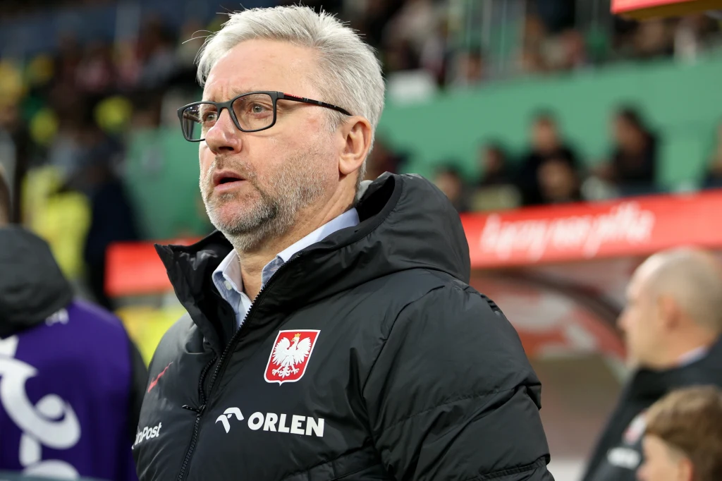 Mężczyzna w okularach i z siwymi włosami stoi w kurtce z godłem Polski i napisem Orlen na tle stadionu piłkarskiego, w tle widoczne rozmyte sylwetki oraz fragment czerwonego banneru.