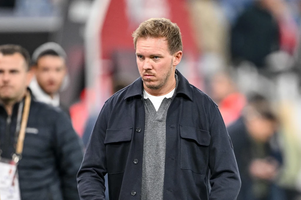 Julian Nagelsmann Mężczyzna o jasnych włosach ubrany w ciemną kurtkę i szary sweter stoi na pierwszym planie, w tle widoczne rozmyte postacie osób.