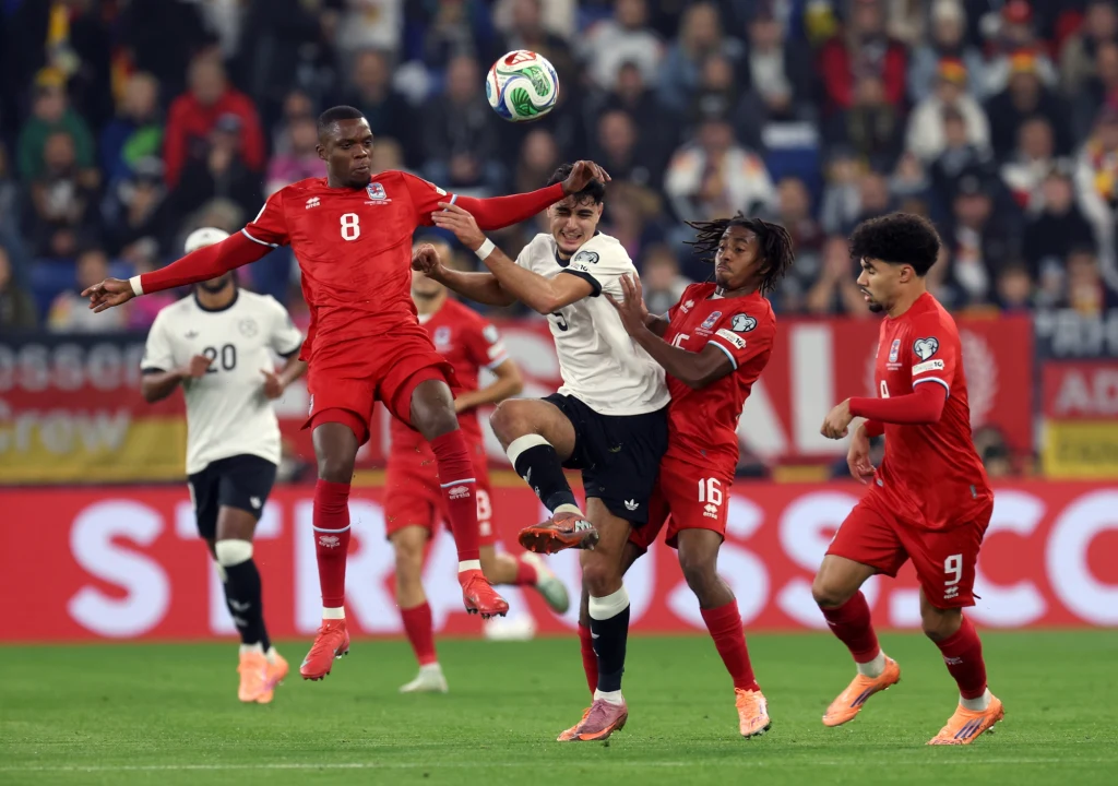 Kadr z meczu Niemcy - Luksemburg Zacięta walka o piłkę podczas meczu piłki nożnej, trzech zawodników w czerwonych strojach rywalizuje z jednym piłkarzem w białej koszulce, wokół nich widać tłum obserwujących kibiców.