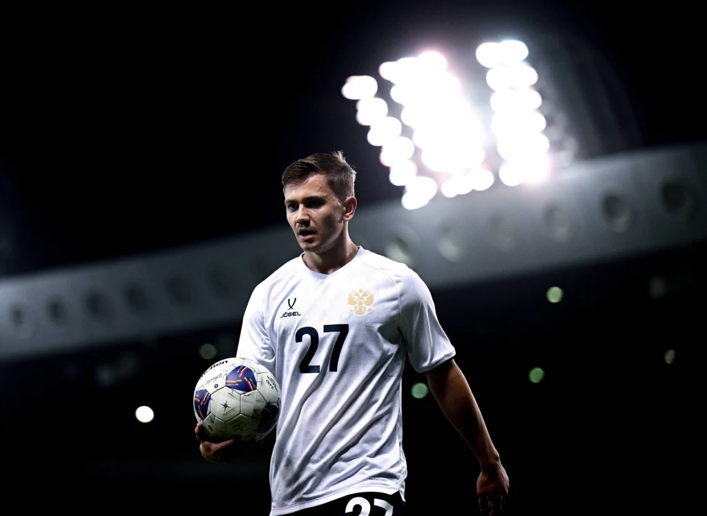 Piłkarz w białym stroju z numerem 27 trzyma piłkę na tle jasno oświetlonego stadionu, wyrazista gra świateł podkreśla atmosferę wieczornego meczu.